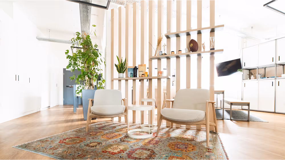 Our Fürther entrance area with two beige armchairs, small round tables, a patterned rug, and wooden slat room divider with plants and decorative items