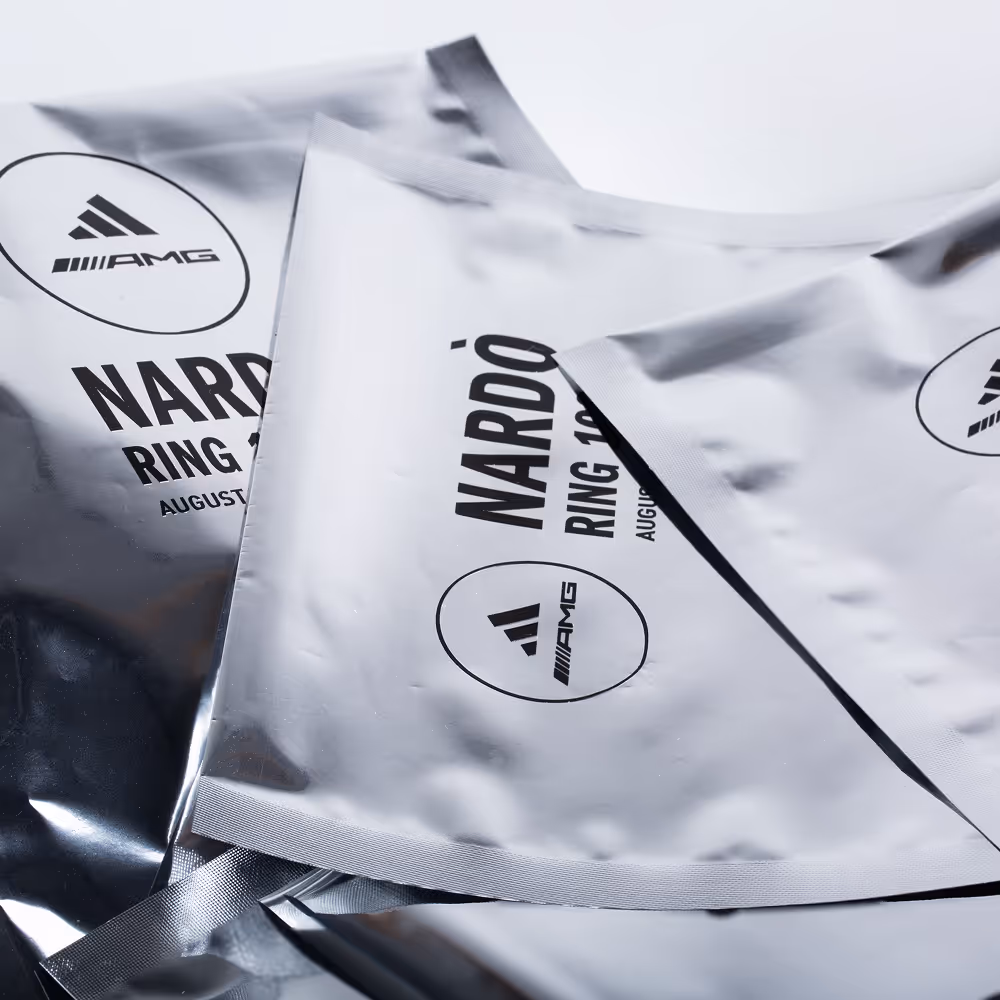 Close-up of silver glossy packages printed with adidas and AMG logos and the text ‘Nardò Ring 100km’.