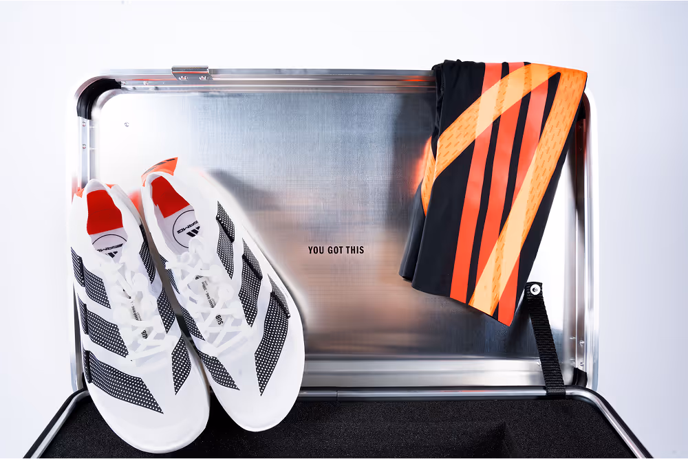 Open metal case with foam insert holding white sports shoes with black stripes and red details, plus a black garment with orange stripes. The lid reads ‘YOU GOT THIS’.
