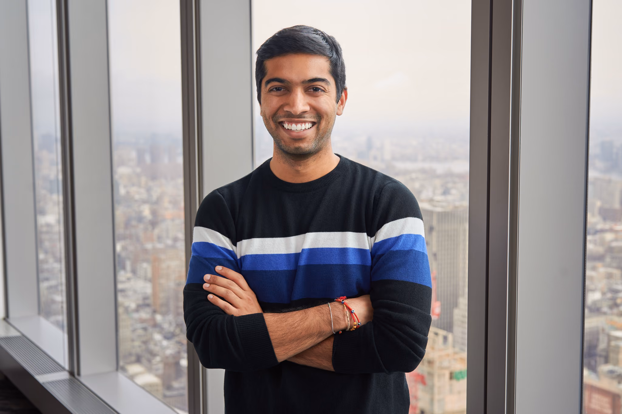 Smiling man with crossed arms in black sweater with blue and white stripes by a tall office window.