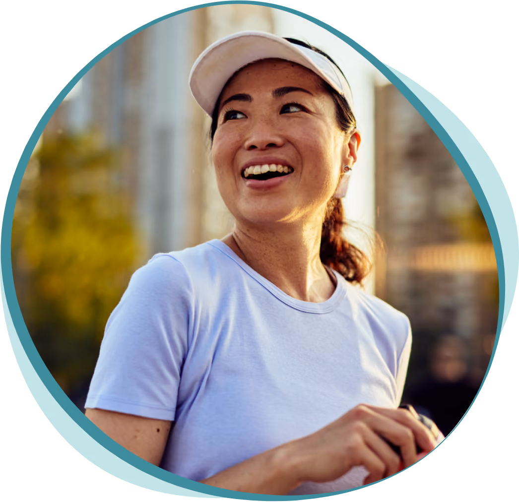 Smiling woman in a white visor and light purple shirt jogging outdoors in an urban setting at sunset.
