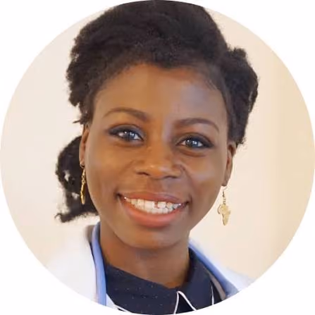 Smiling woman with natural hair wearing gold Africa-shaped earrings and a white coat over a dark top.