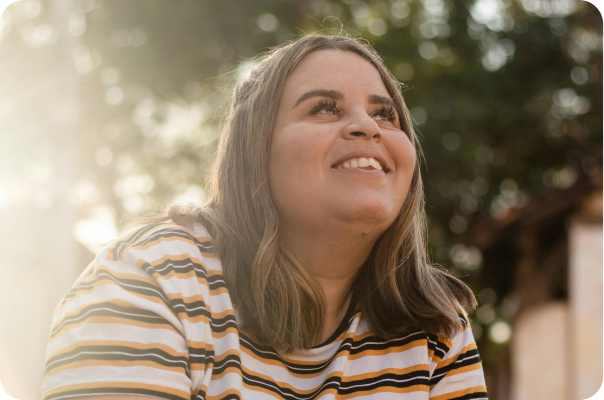 Smiling woman with shoulder-length brown hair wearing a white, black, and yellow striped shirt looking upwards, with sunlight and greenery in the background.