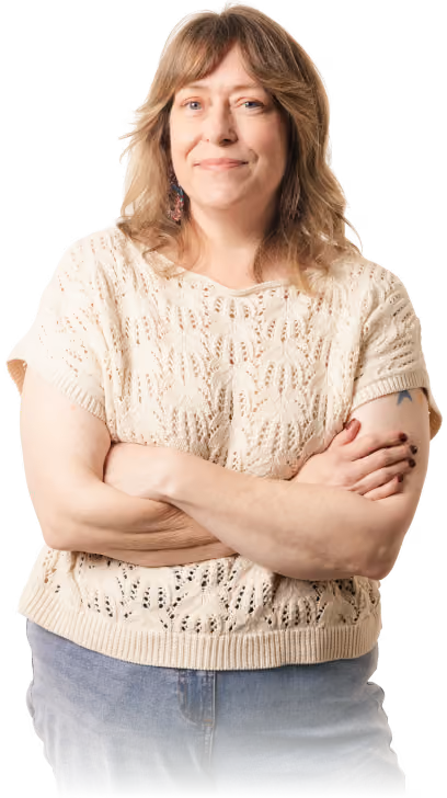 A middle-aged woman with shoulder-length light brown hair, wearing a cream knit short-sleeve sweater and blue jeans, stands with arms crossed and a slight smile.