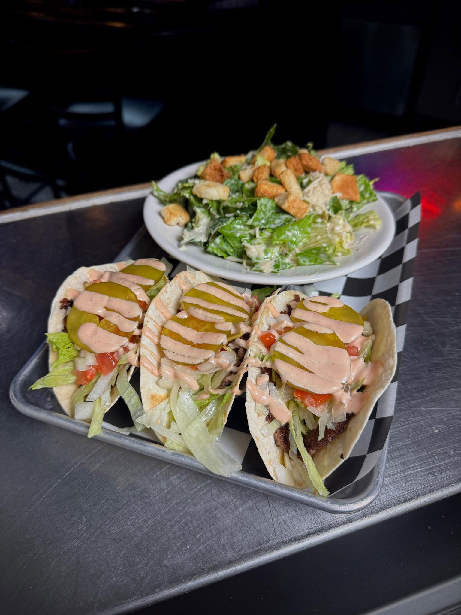 Three metal trays with buffalo chicken wings, celery sticks, carrot sticks, and dipping sauce on a stainless steel table.