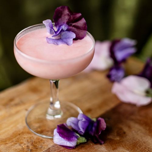 A beautiful pink craft cocktail with bright purple edible flower garnishes from event bartenders at Tuxedos & Tennis Shoes Catering, a DSquared Hospitality Company in Seattle