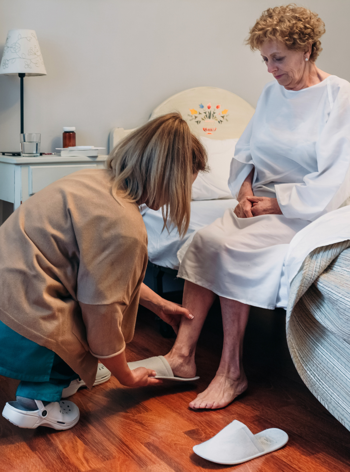 A caregiver gently assists an elderly woman with her slippers in a cozy, well-lit bedroom, emphasizing personalized and caring assistance.