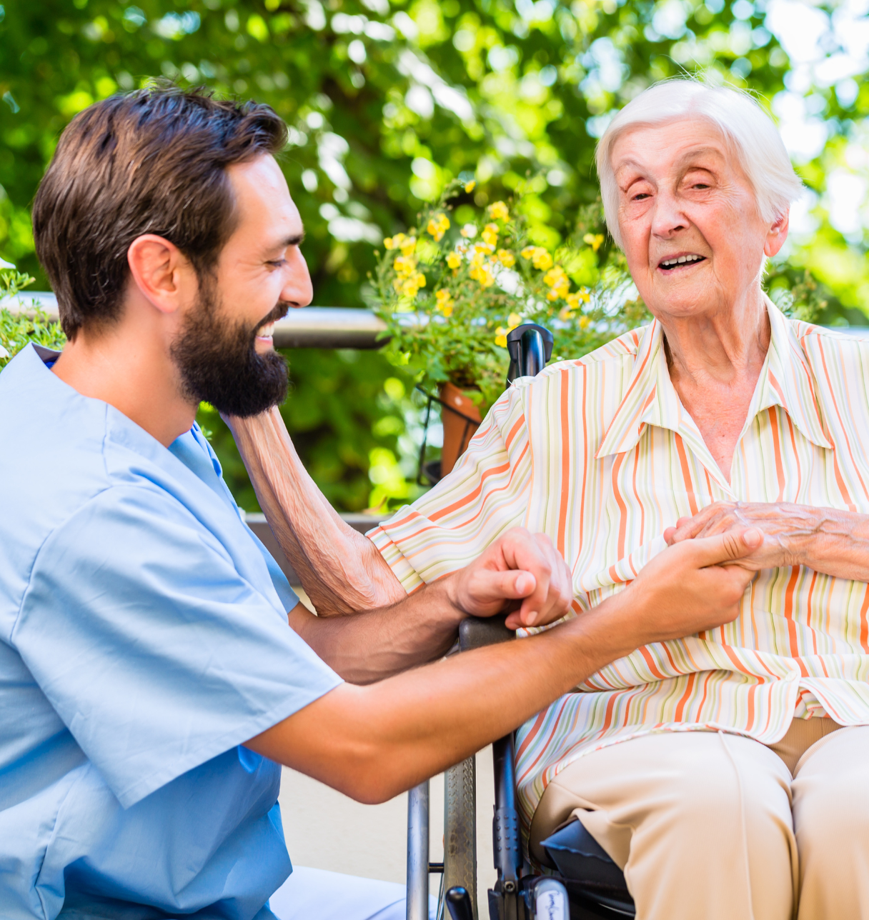 An elderly woman in a striped blouse enjoys a conversation with a smiling male caregiver in a blue uniform outdoors, surrounded by greenery.