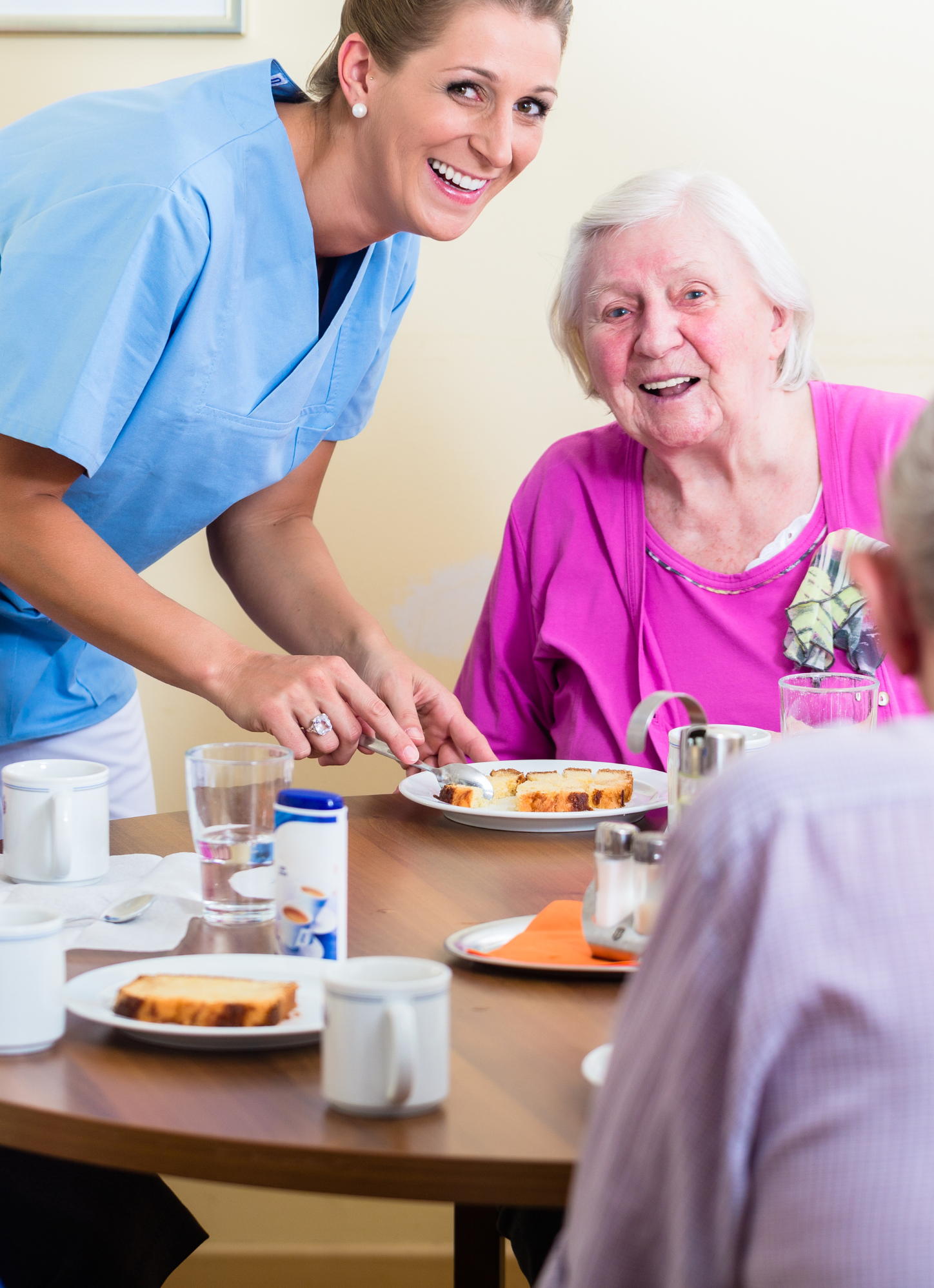 A caregiver in a blue uniform serves food to an elderly woman in pink, who is seated at a dining table, enhancing her mealtime experience.