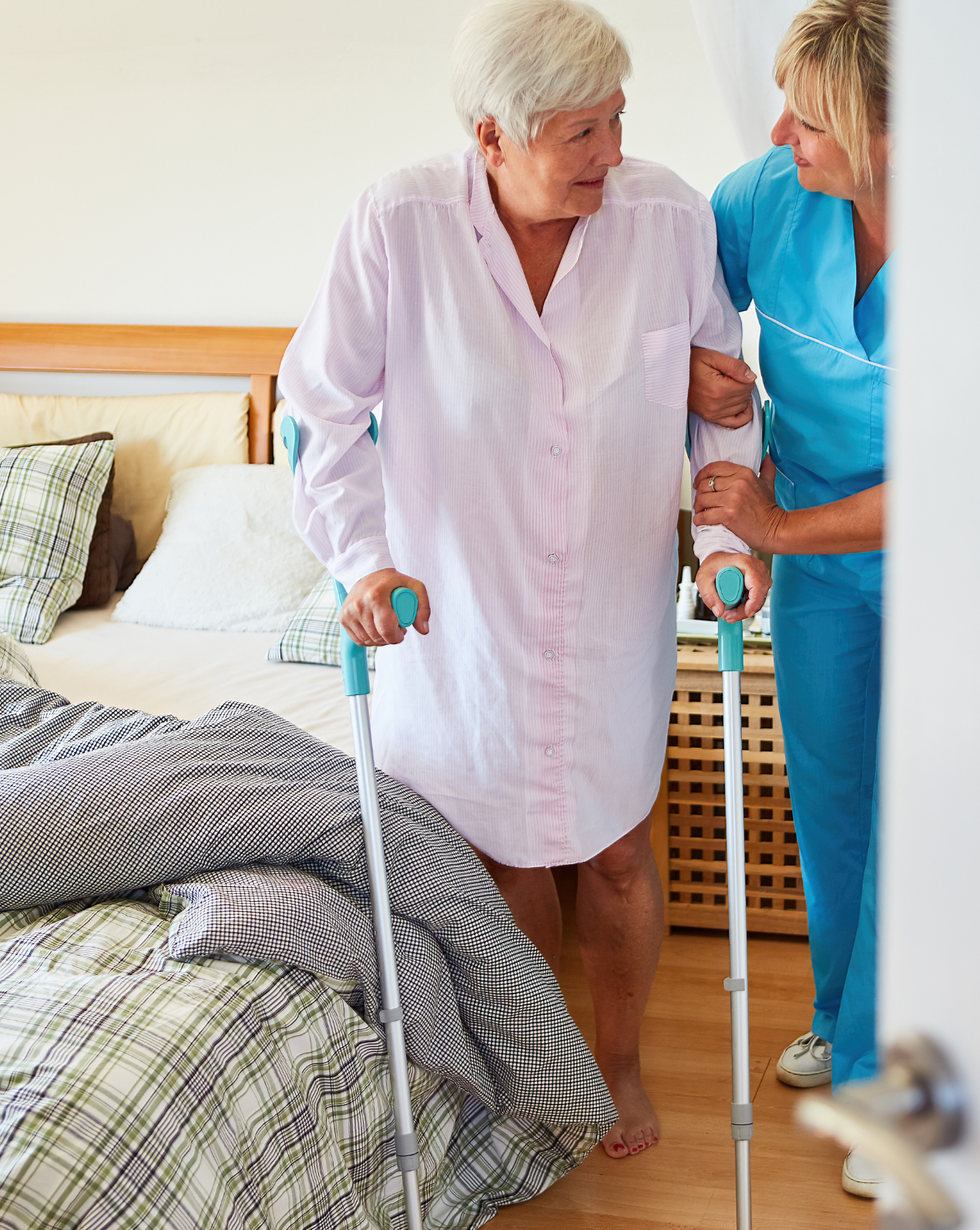 A caregiver in a teal uniform supports an elderly woman using a walker in her bedroom, providing stability and care.