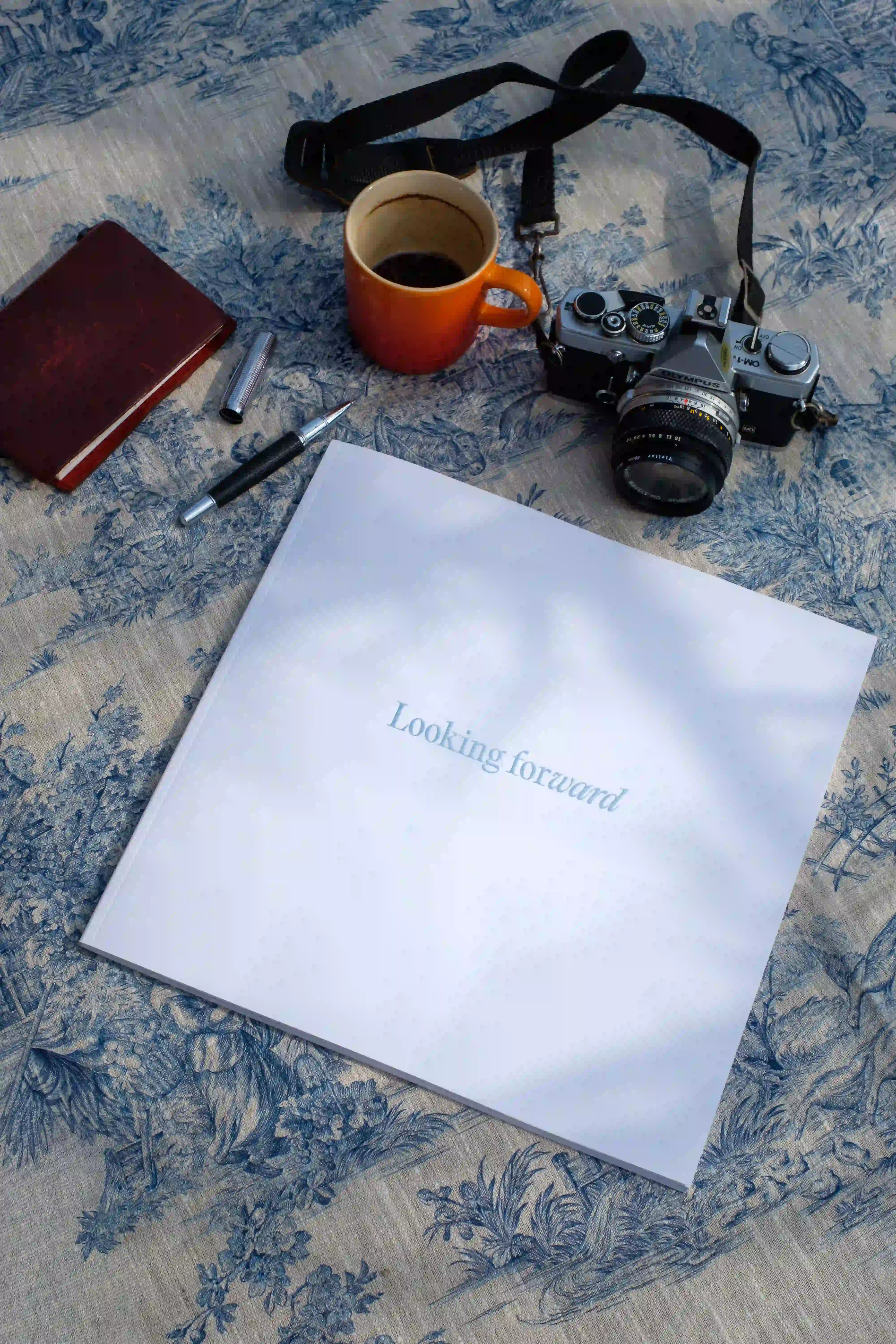 Publication titled “Looking forward” placed on a patterned tablecloth beside a camera, notebook, pen and coffee cup.