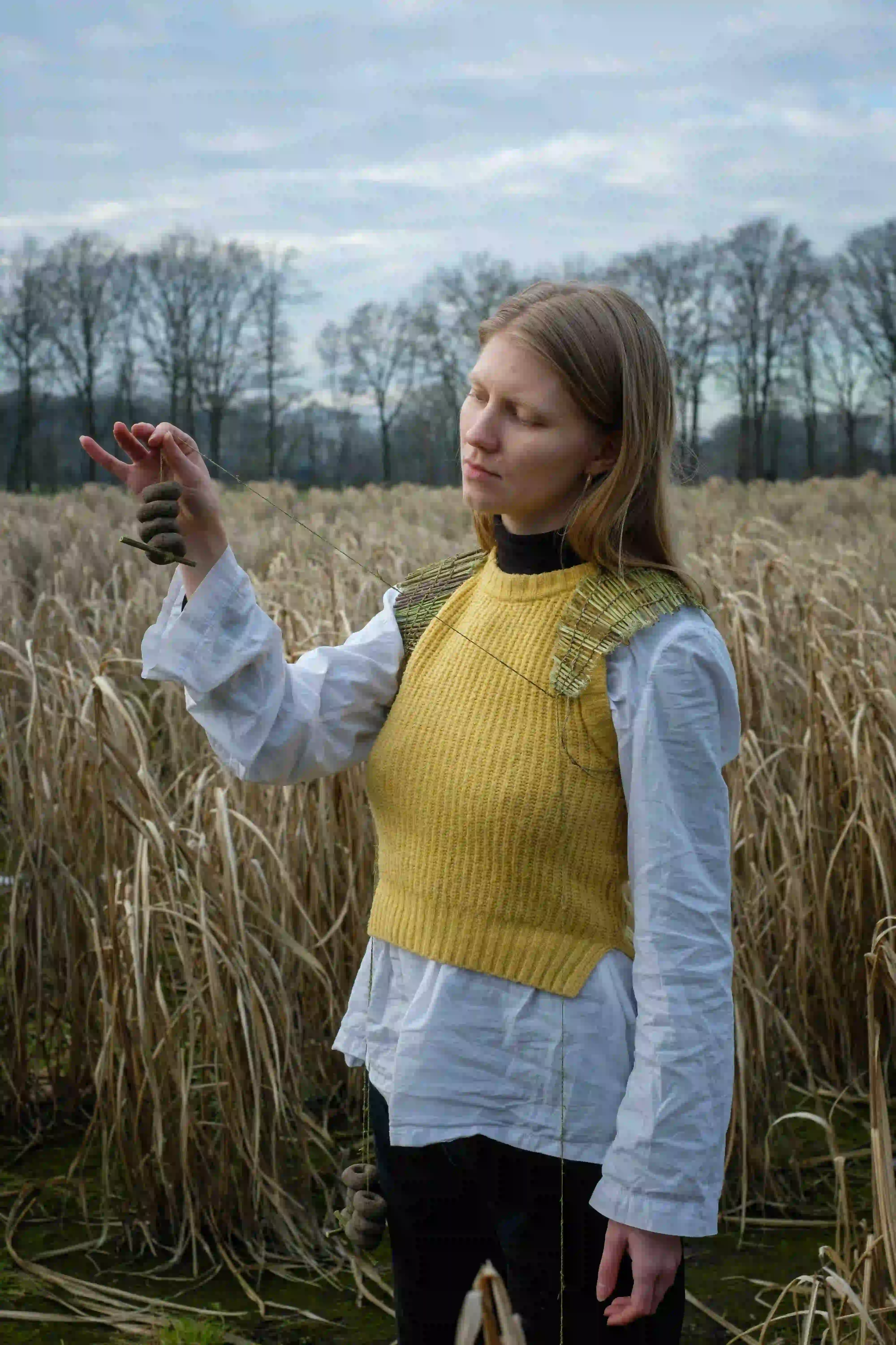 Person standing in a field holding a string with clay forms, wearing a yellow knitted vest.