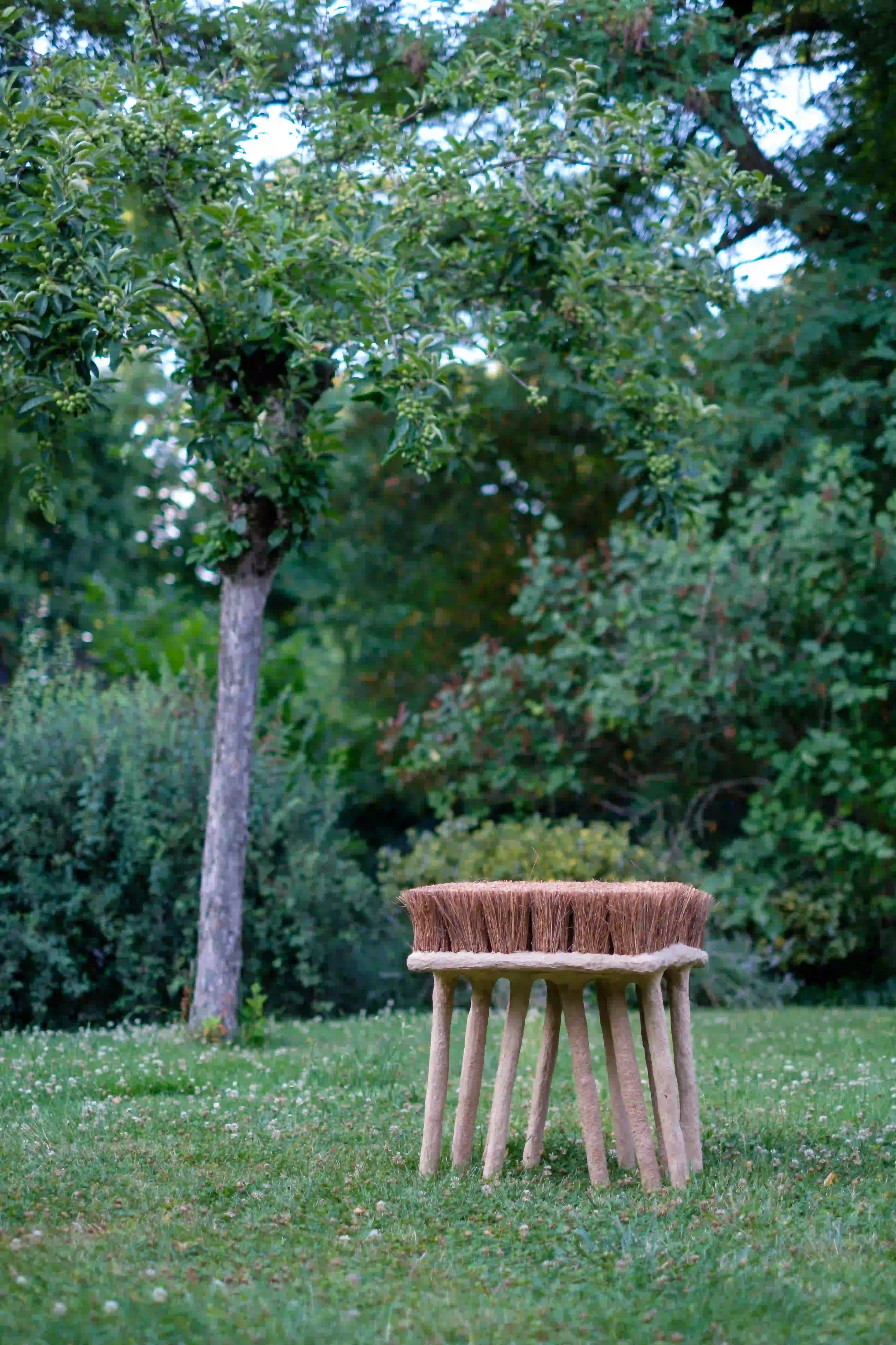 Handmade stool with broom-like bristles and wooden legs placed on grass in a garden.