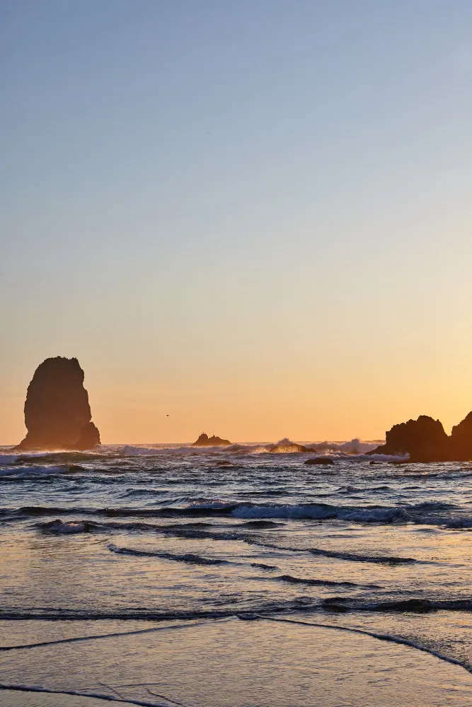 Sunset over Cannon Beach near Portland, Oregon, with gentle waves reflecting the warm orange glow and sea stacks rising from the ocean.