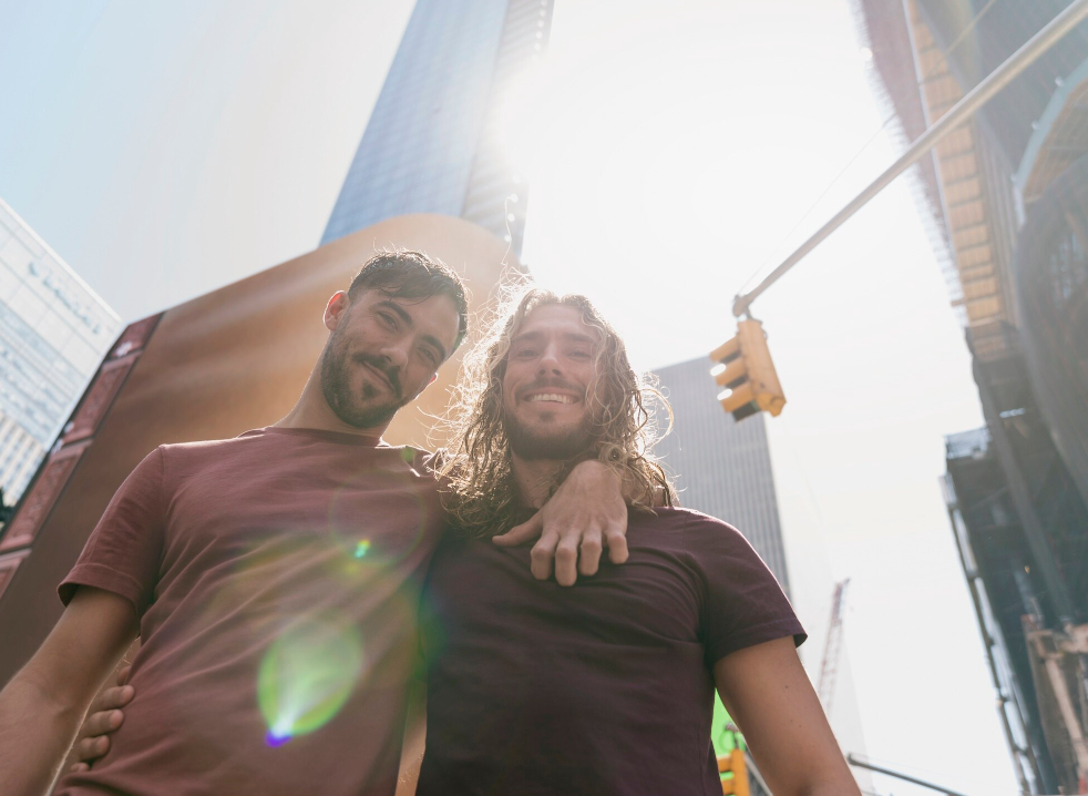 Two men stand close together smiling in bright sunlight with city buildings behind them, symbolizing hope, connection, and recovery after addiction treatment