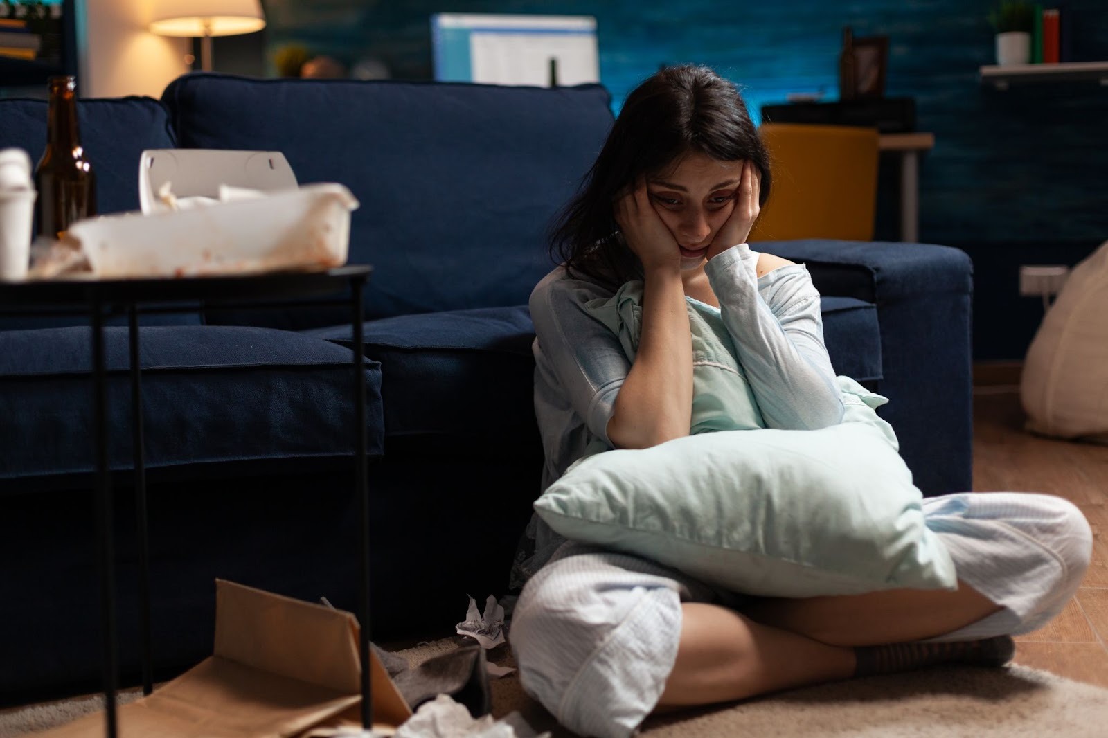 A woman sits on the floor in front of a couch at night, holding a pillow and looking distressed. There are beer bottles, food containers, and tissues around her.