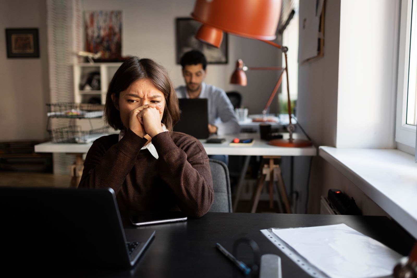 A woman sitting on a desk in front of a laptop visibly stressed, with an office setting backdrop