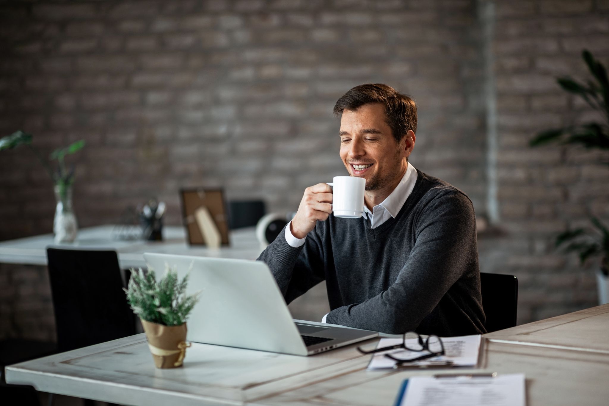 A man sitting at a desk with a laptop, smiling while drinking coffee, representing balancing daily life with outpatient treatment
