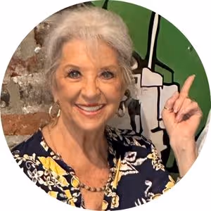 Smiling elderly woman with gray hair pointing upwards against a green and brick background.