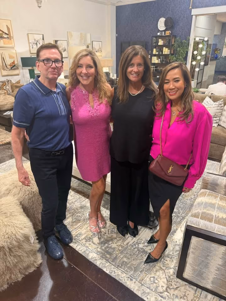 A man wearing glasses and a navy blue polo shirt stands next to three women posing indoors; one woman wears a bright pink dress, another wears a black outfit with a pearl necklace, and the third wears a hot pink blouse with a black skirt and a burgundy Yves Saint Laurent purse, all standing on a patterned rug in a warmly decorated living room.