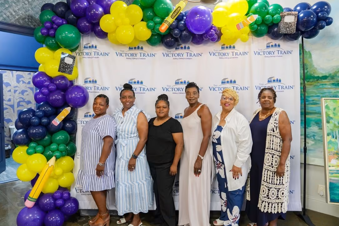 Six women standing side by side in front of a backdrop with The Victory Train logo, framed by a colorful balloon arch in green, yellow, purple, and blue with pencil and notebook decorations.