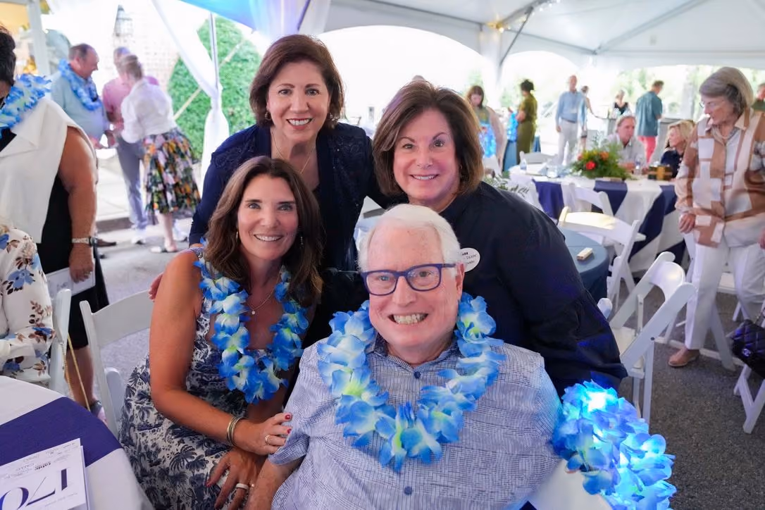 Four adults smiling and posing for a photo under a white event tent, three women standing behind a seated man who wears glasses and a blue floral lei, with white folding chairs and tables with decorations visible in the background.