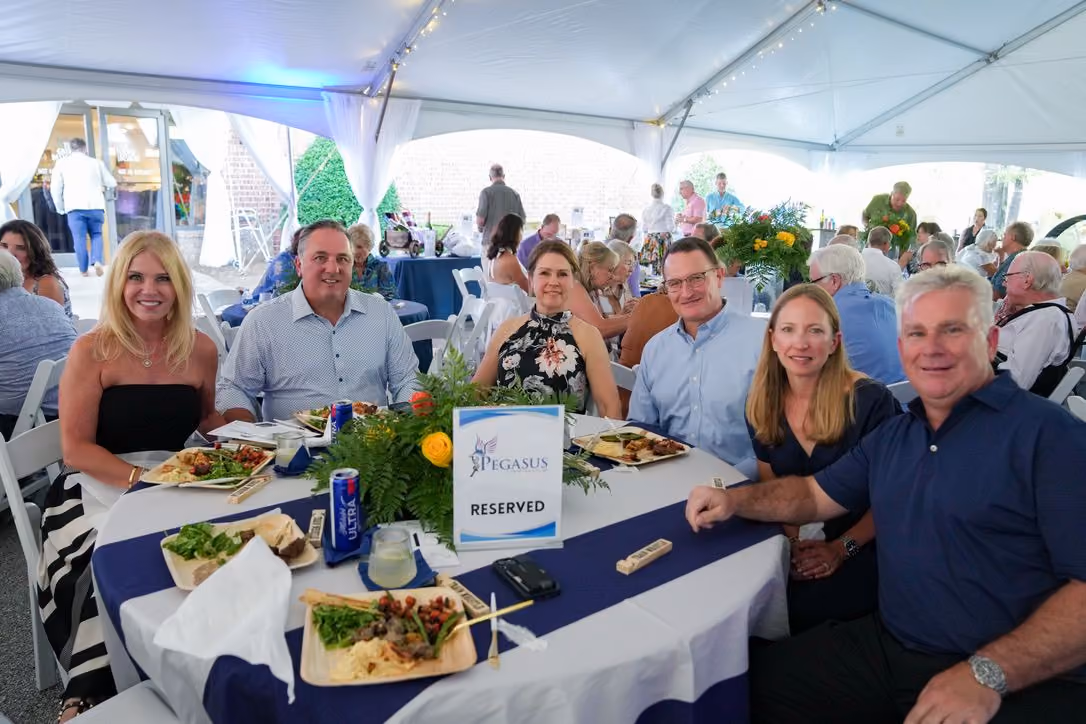 Group of six adults seated around a white table with a blue runner under a large white event tent, enjoying a meal with plates of food, drinks, and a centerpiece with flowers and a reserved sign labeled 'Pegasus'.