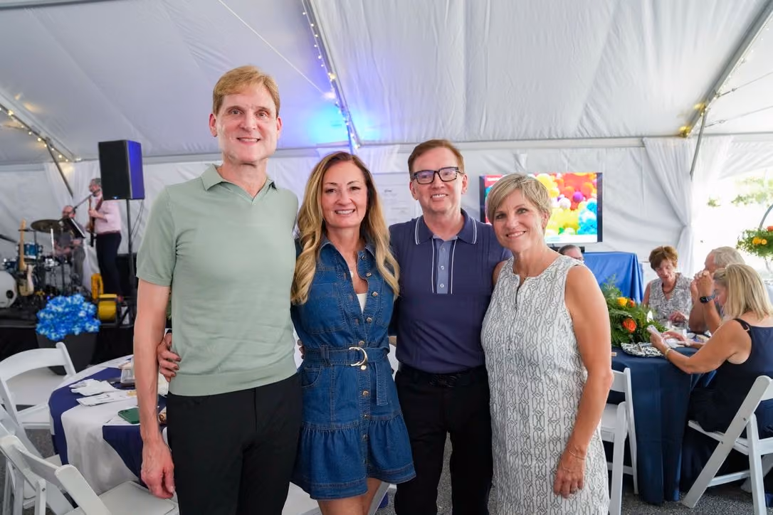 Four adults standing closely and smiling under a large white event tent, with a stage and musical equipment in the background and people seated at tables.