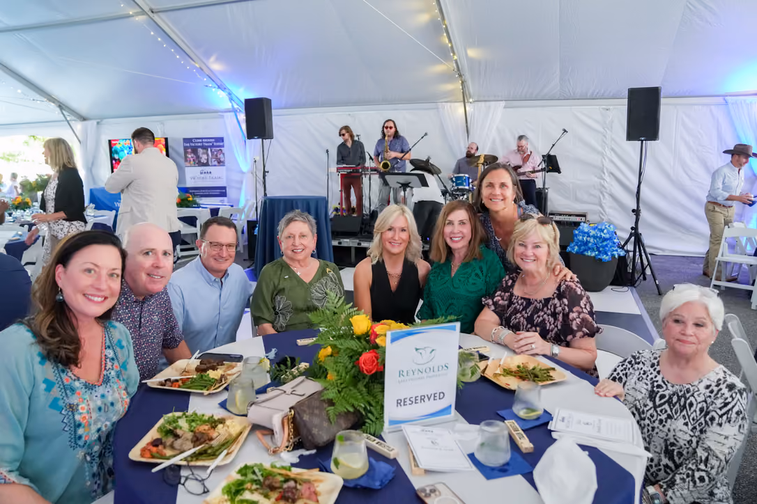 A group of nine adults sitting around a round table with plates of food in a large white event tent, with a live band playing on a stage in the background and a sign on the table that reads 'Reynolds Medical Center RESERVED.'