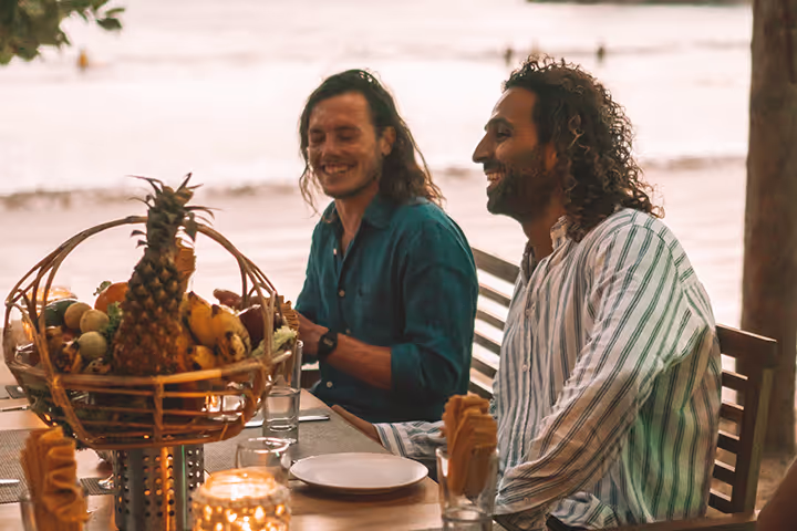 Two men sitting at a table with a basket of fruit and a pineapple.