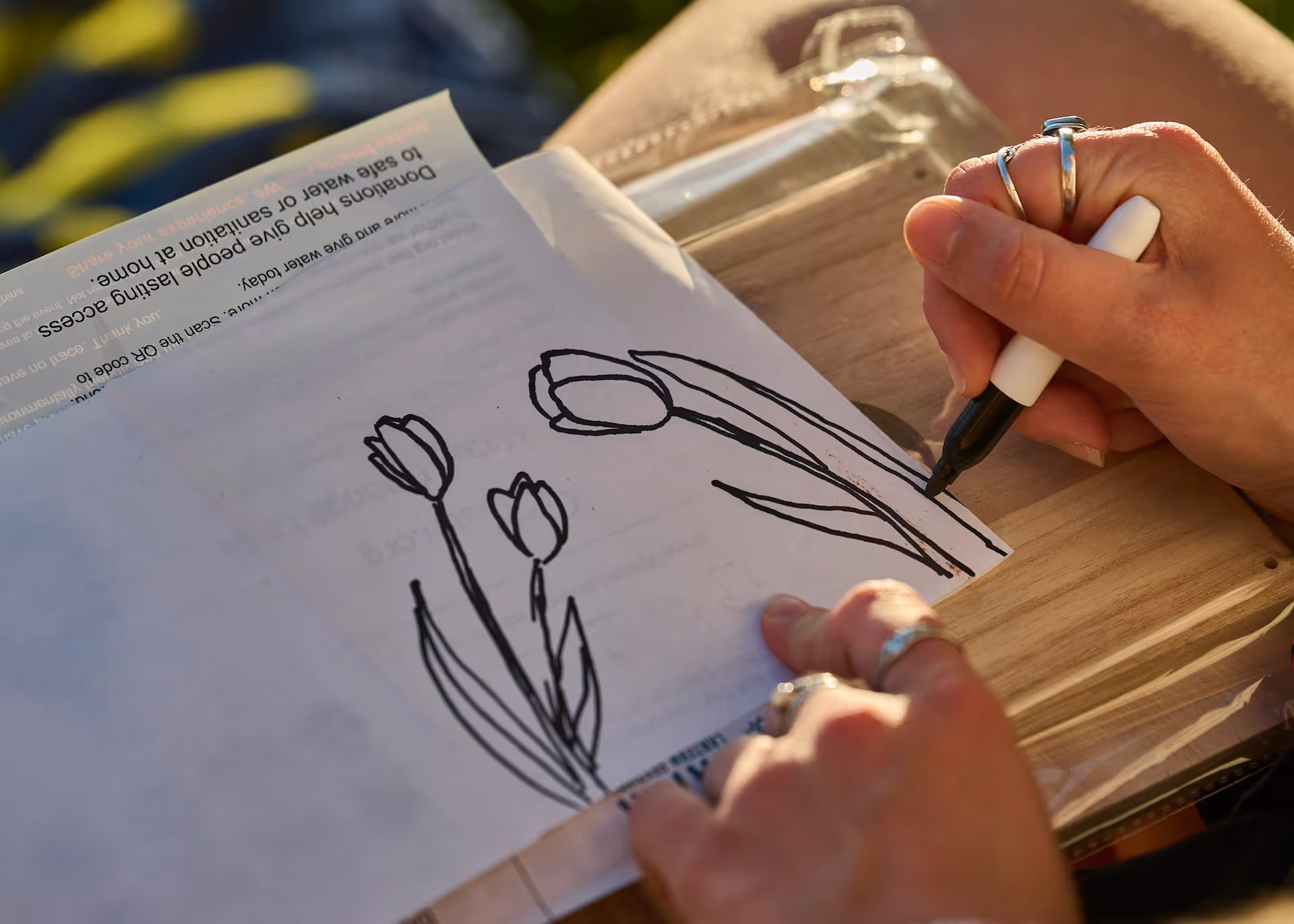 Person drawing black outline of tulip flowers on white paper using a black marker, with sunlight casting shadows.