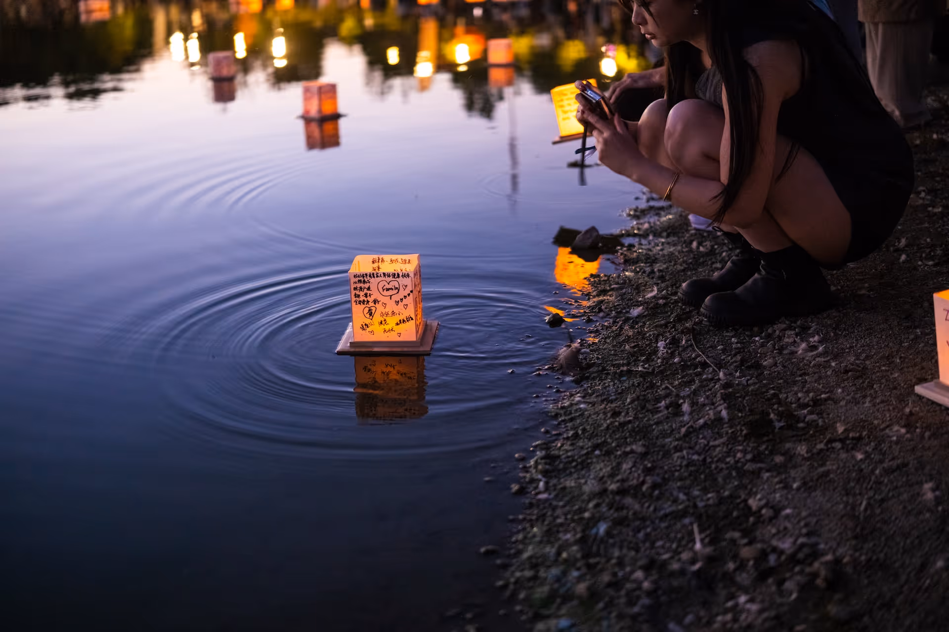 A woman crouching by a lakeshore at dusk, taking a photo of illuminated floating paper lanterns on the water.