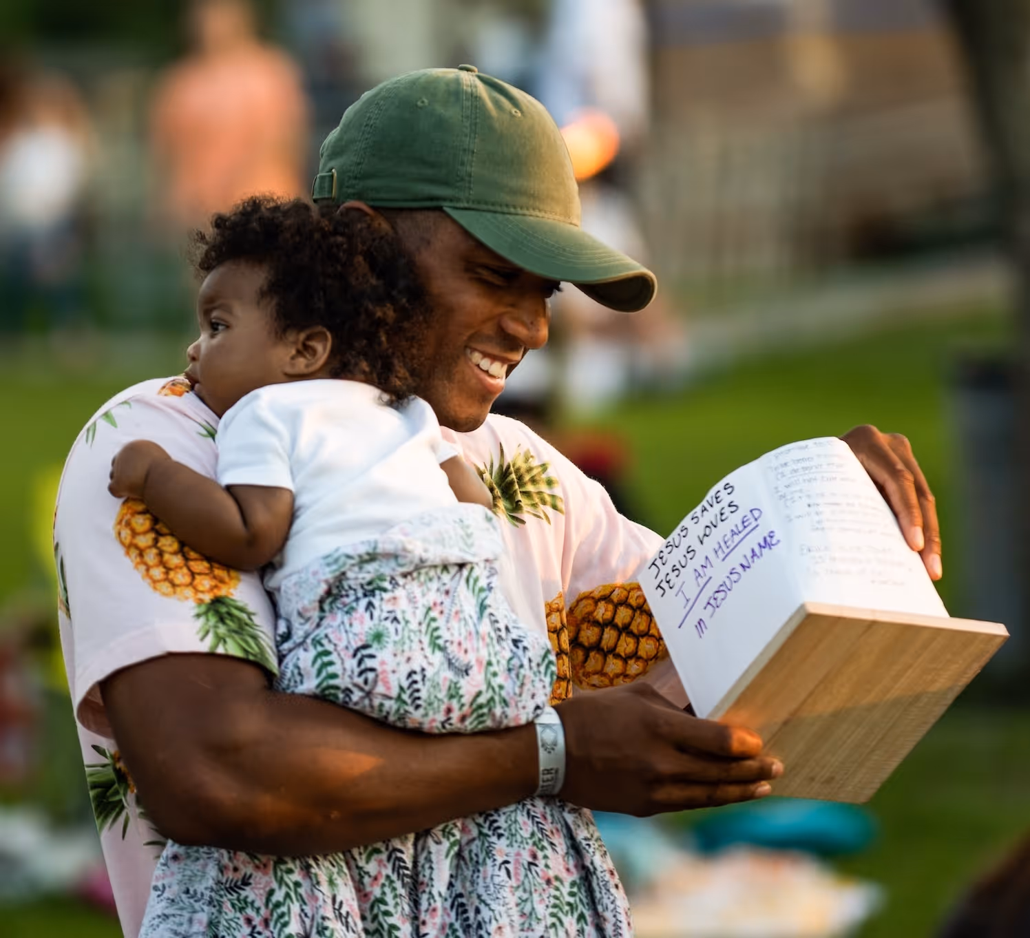 Smiling man in a green cap holding a child and a book with handwritten religious notes outdoors.