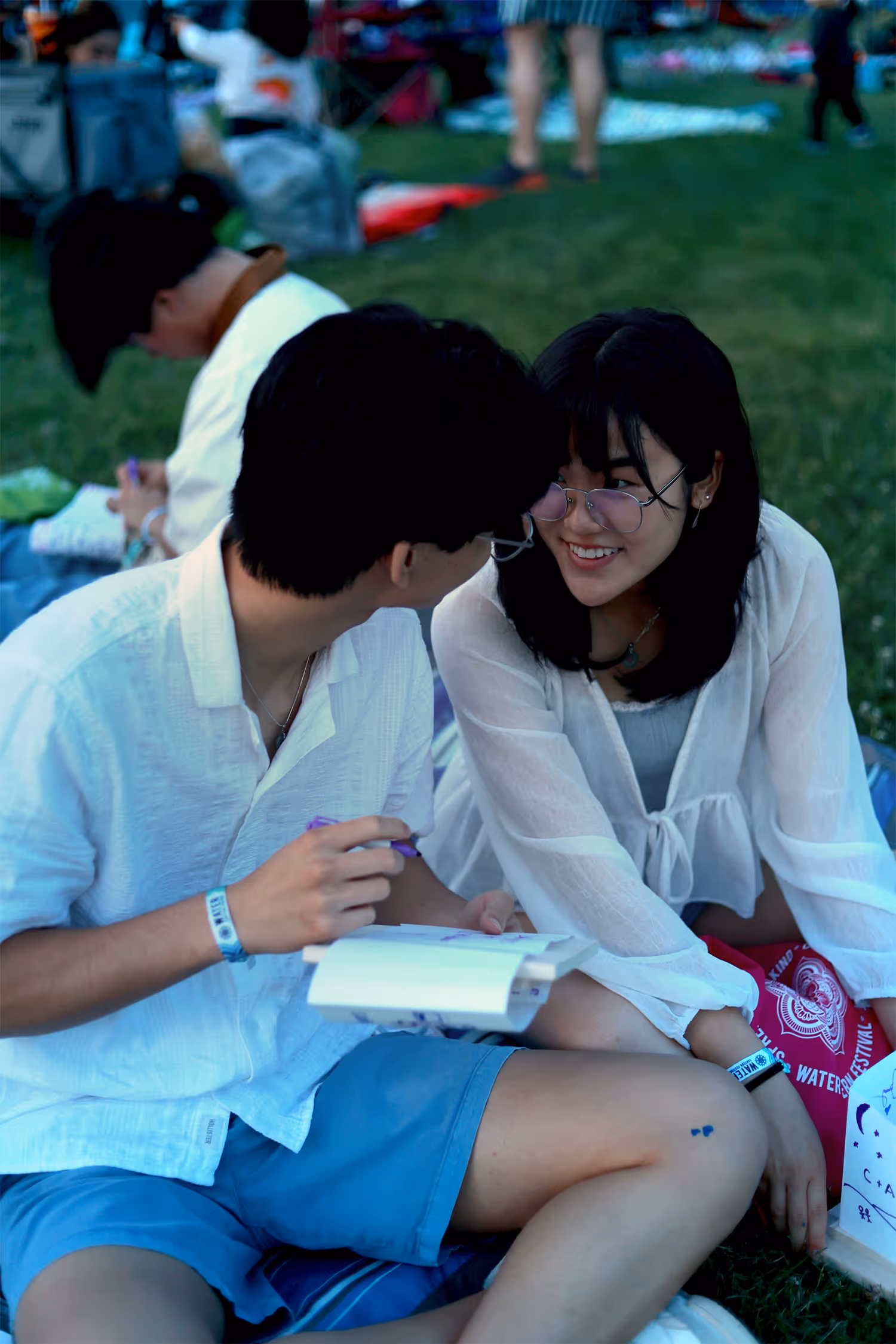 Young man and woman sitting on grass, smiling and talking while holding a notebook and pen.