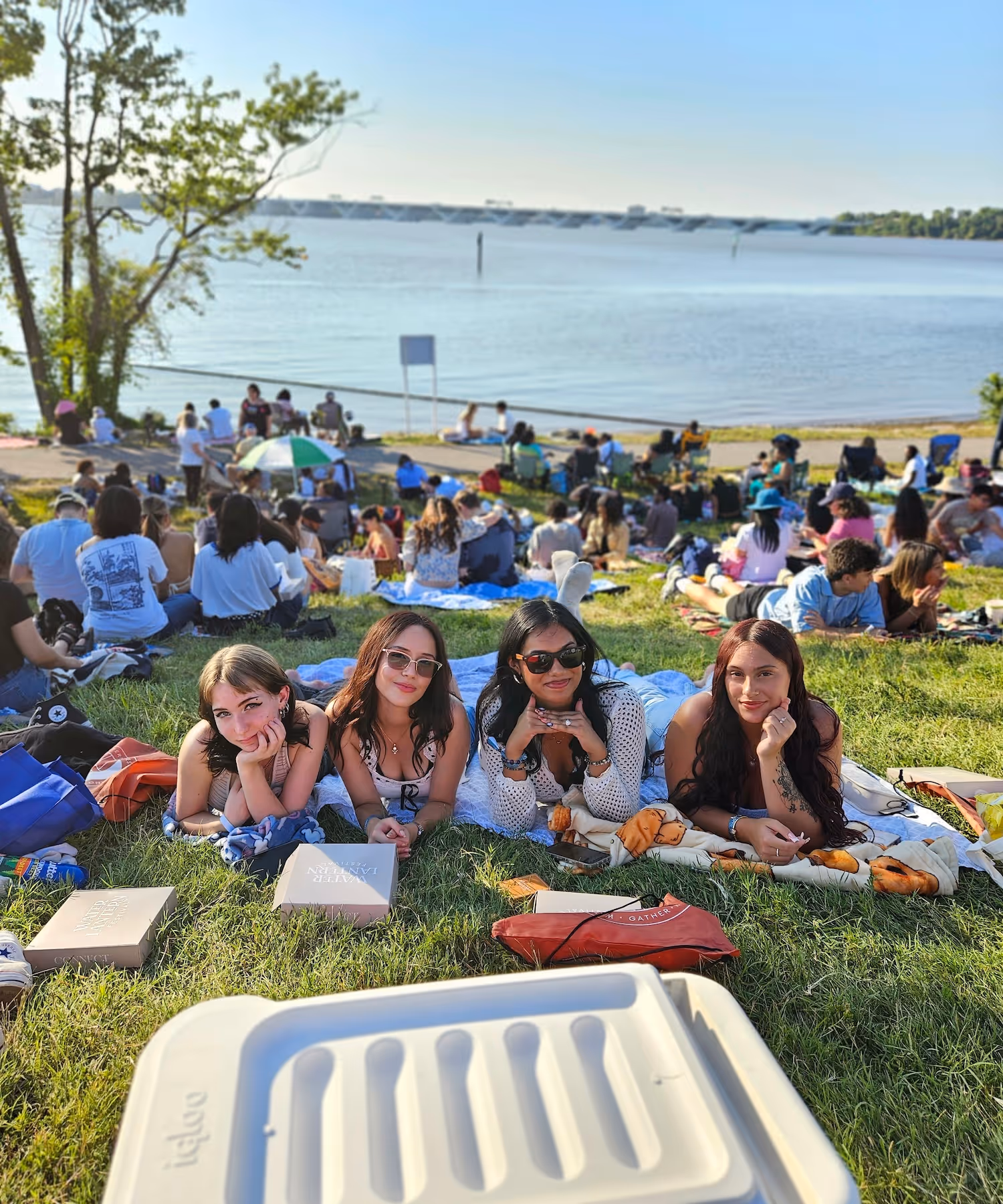 Four young women lying on blankets on grass at a lakeside picnic with many other people in the background under blue sky.