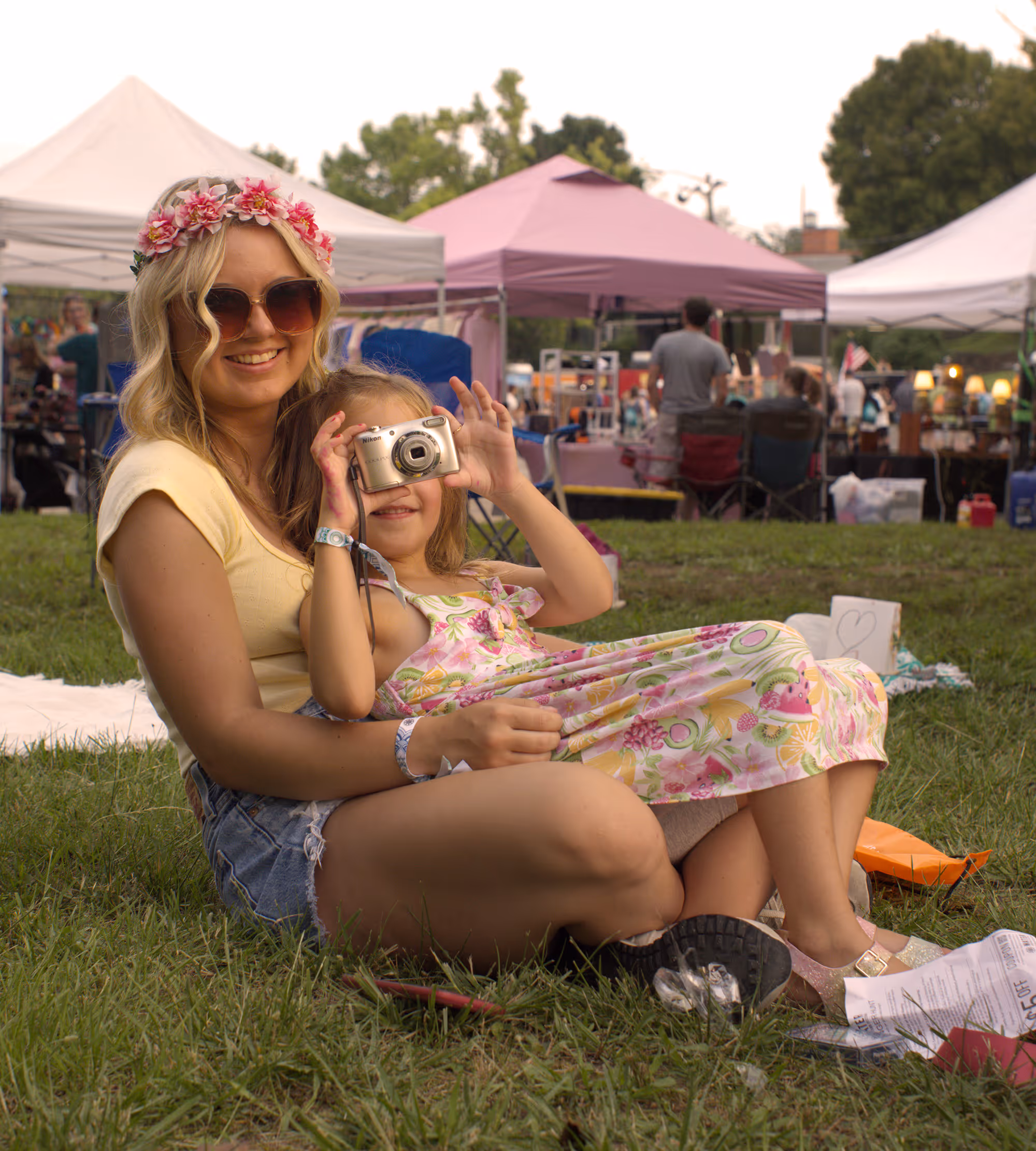 Smiling woman wearing sunglasses and a flower crown sitting on grass with a young girl holding a camera at an outdoor event with tents in the background.