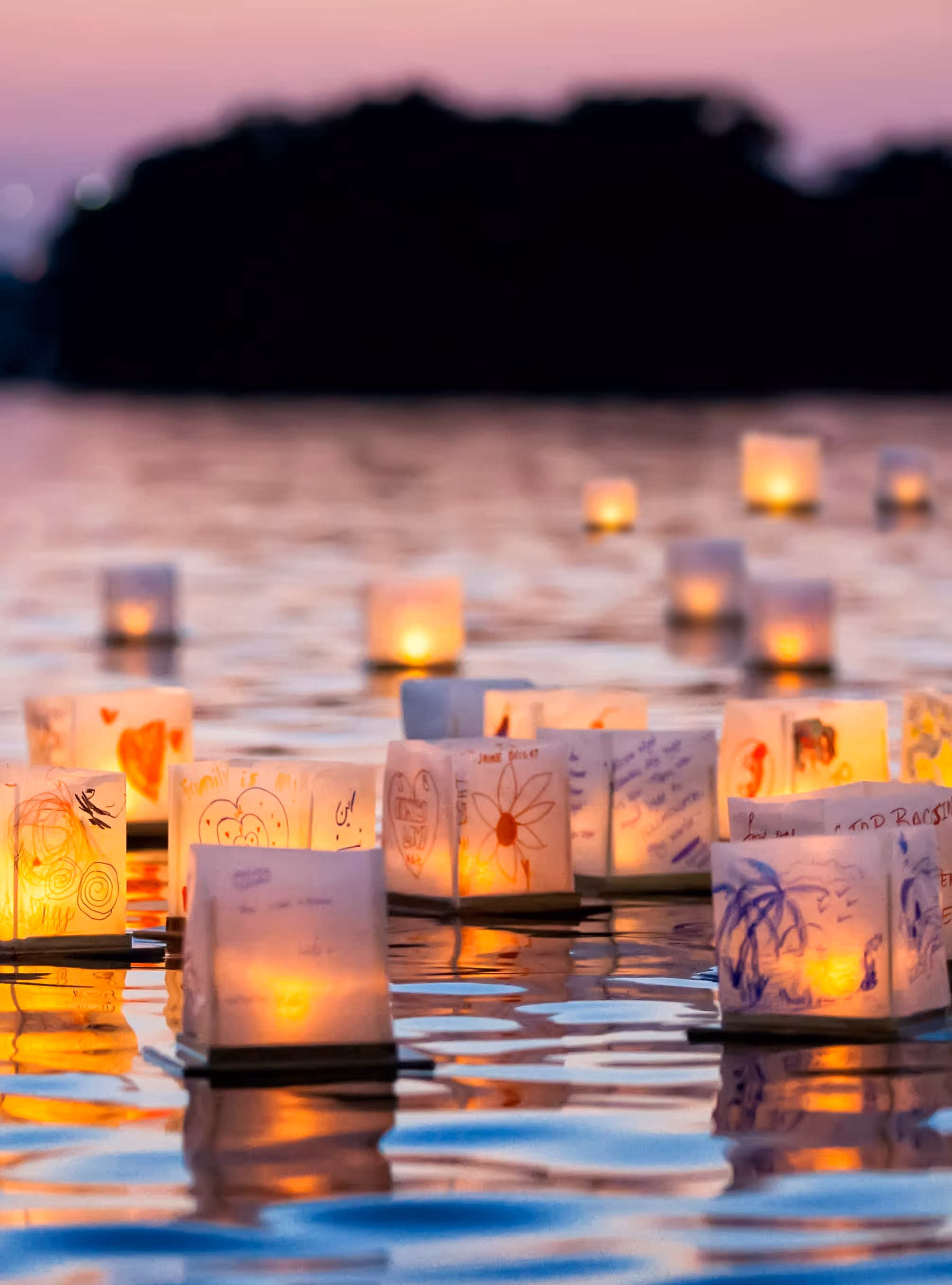 Floating paper lanterns with drawings and candles glowing on calm water at sunset.