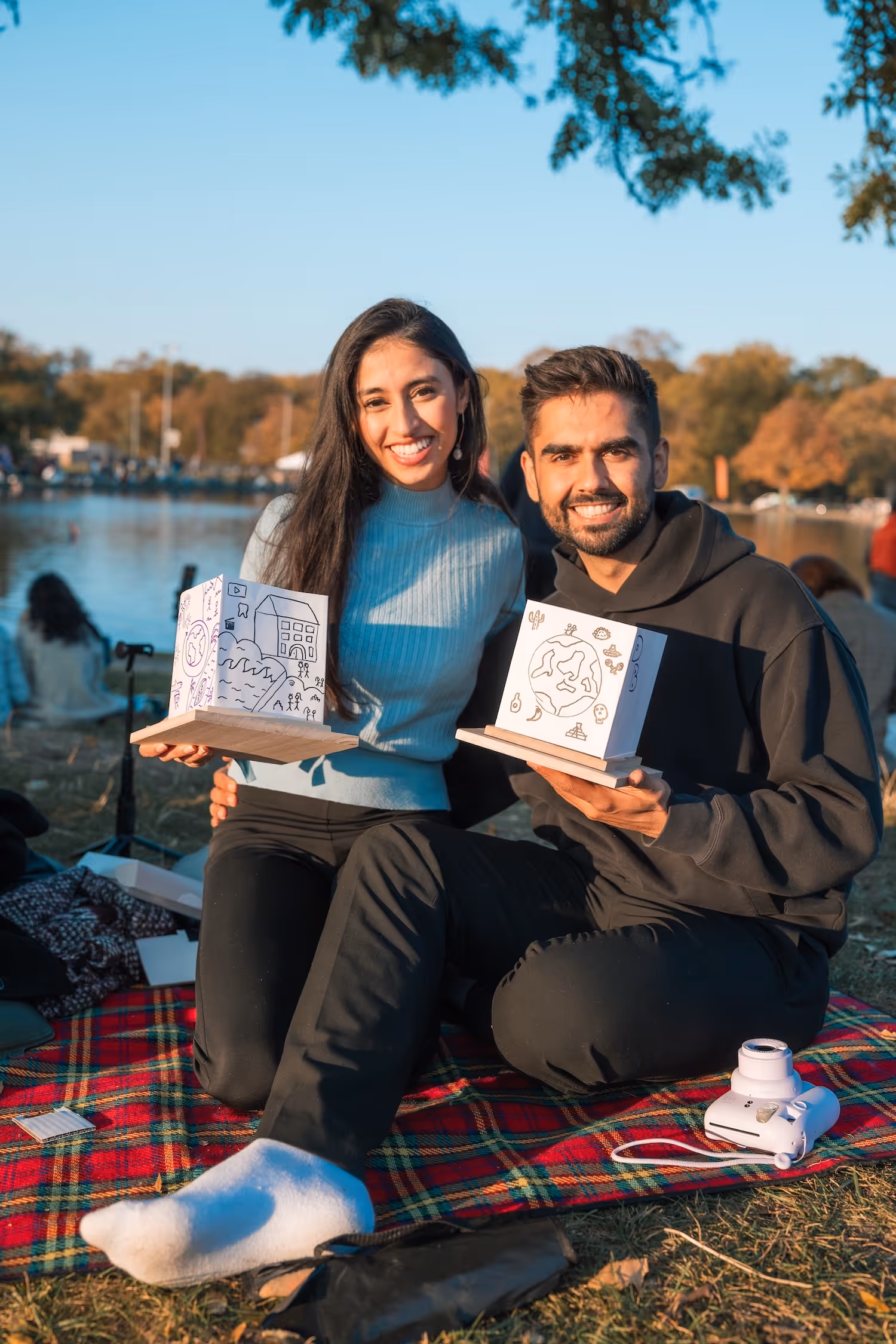 Smiling young couple sitting on a red plaid blanket by a lake, holding handmade paper crafts with drawings.