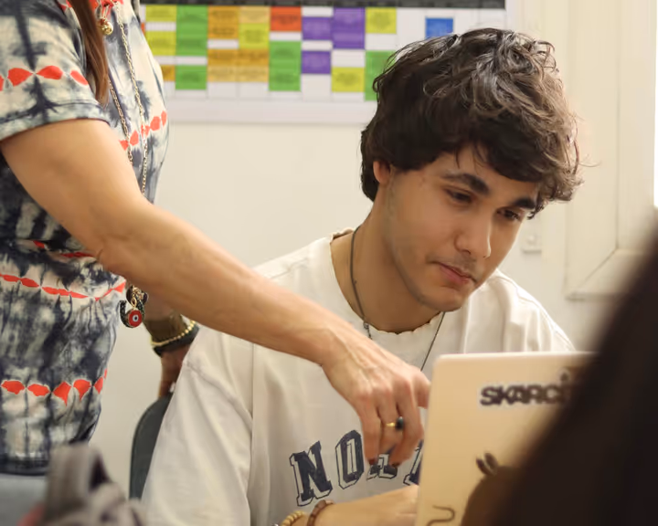 A young man in a white shirt focuses on a laptop screen while a person beside him points at the laptop.