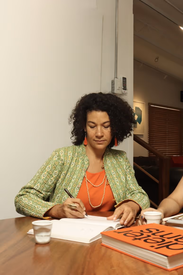 Woman with curly hair in a green patterned jacket and orange top writing in a notebook at a wooden table with coffee cups and an orange book.