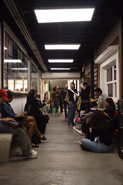 Group of people sitting and standing in a long indoor hallway with fluorescent ceiling lights.