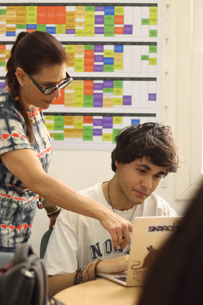 Woman wearing glasses points to a laptop screen while a young man sits at a desk looking at the laptop in a classroom with a colorful schedule on the wall.
