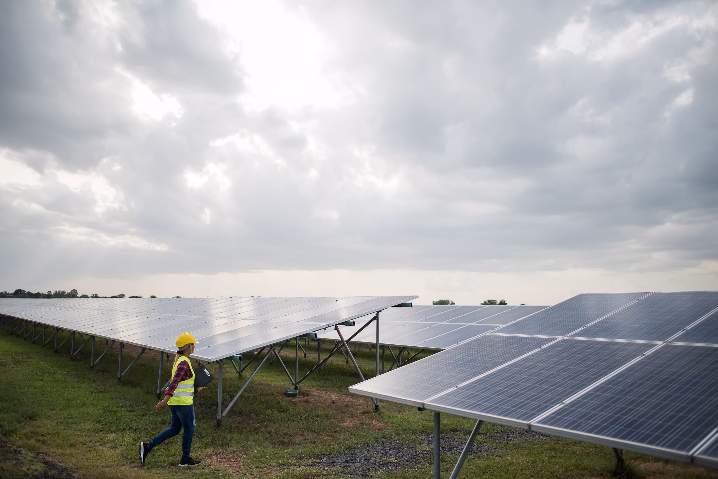 Ingeniera con casco amarillo y chaleco reflectante caminando junto a paneles solares en un campo bajo un cielo nublado.