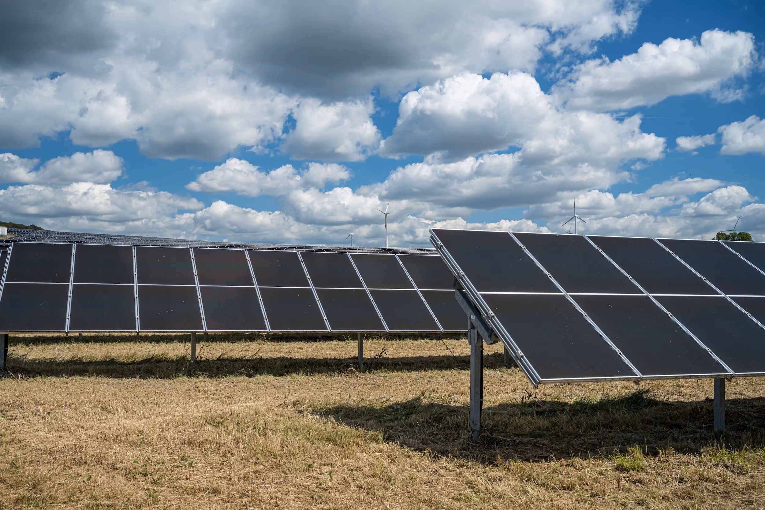 Paneles solares instalados en un campo seco bajo un cielo parcialmente nublado con turbinas eólicas a lo lejos.