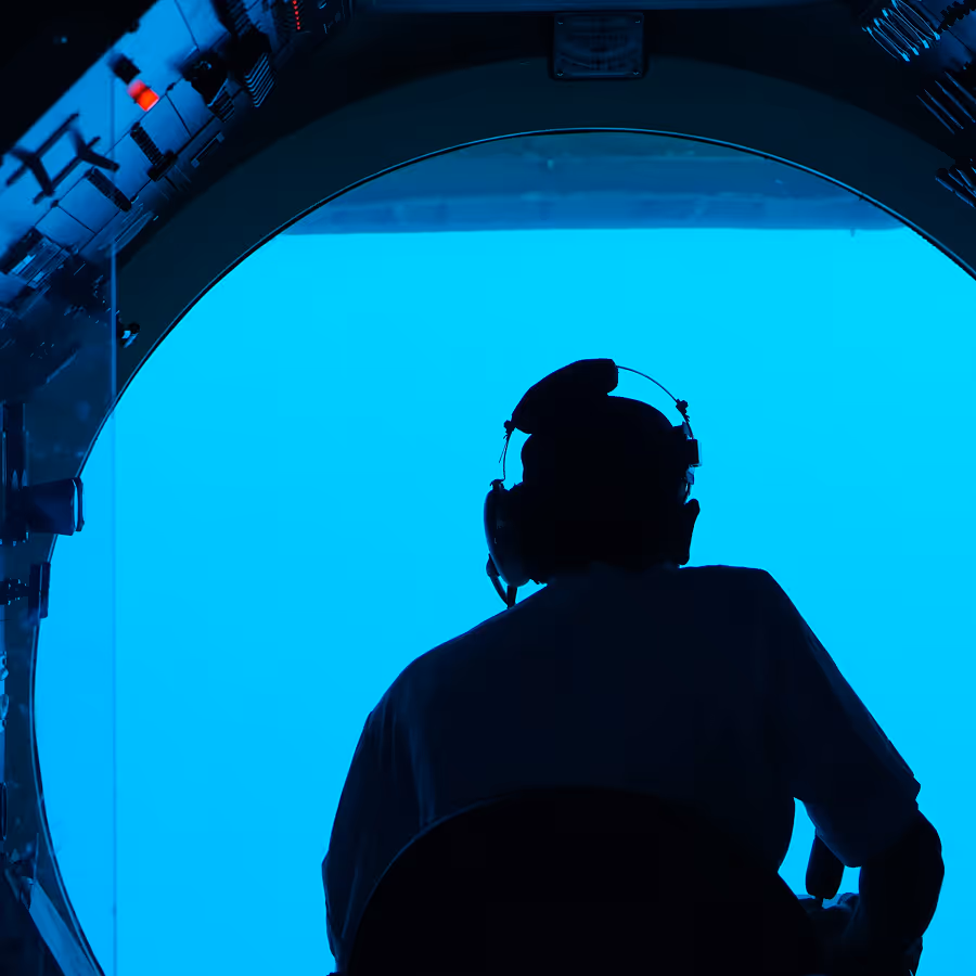 Silhouette of a person wearing a headset inside a submarine window looking out into blue water.