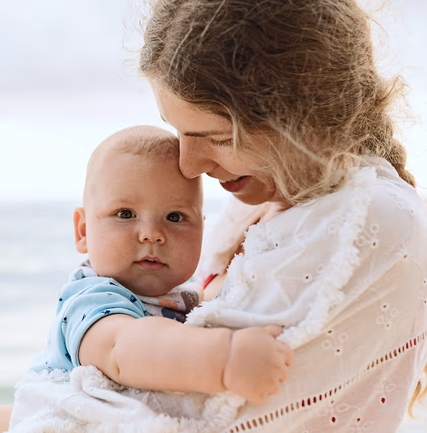 Woman smiling and holding a baby wrapped in a white blanket near the ocean.