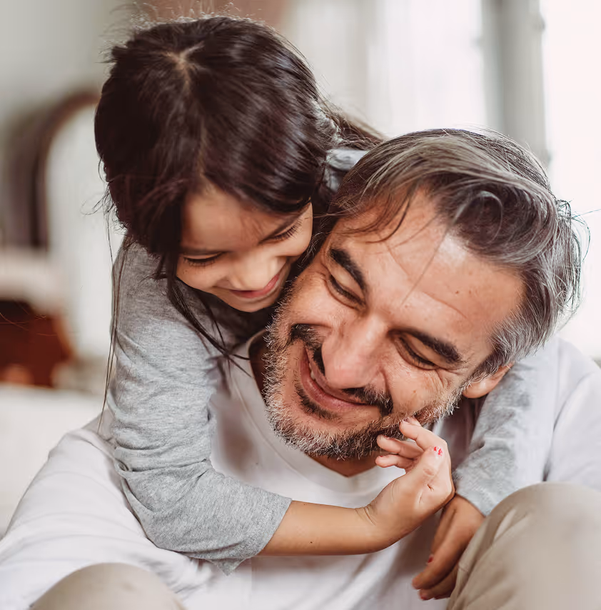 Young girl with dark hair smiling and hugging an older man with gray hair and beard from behind.