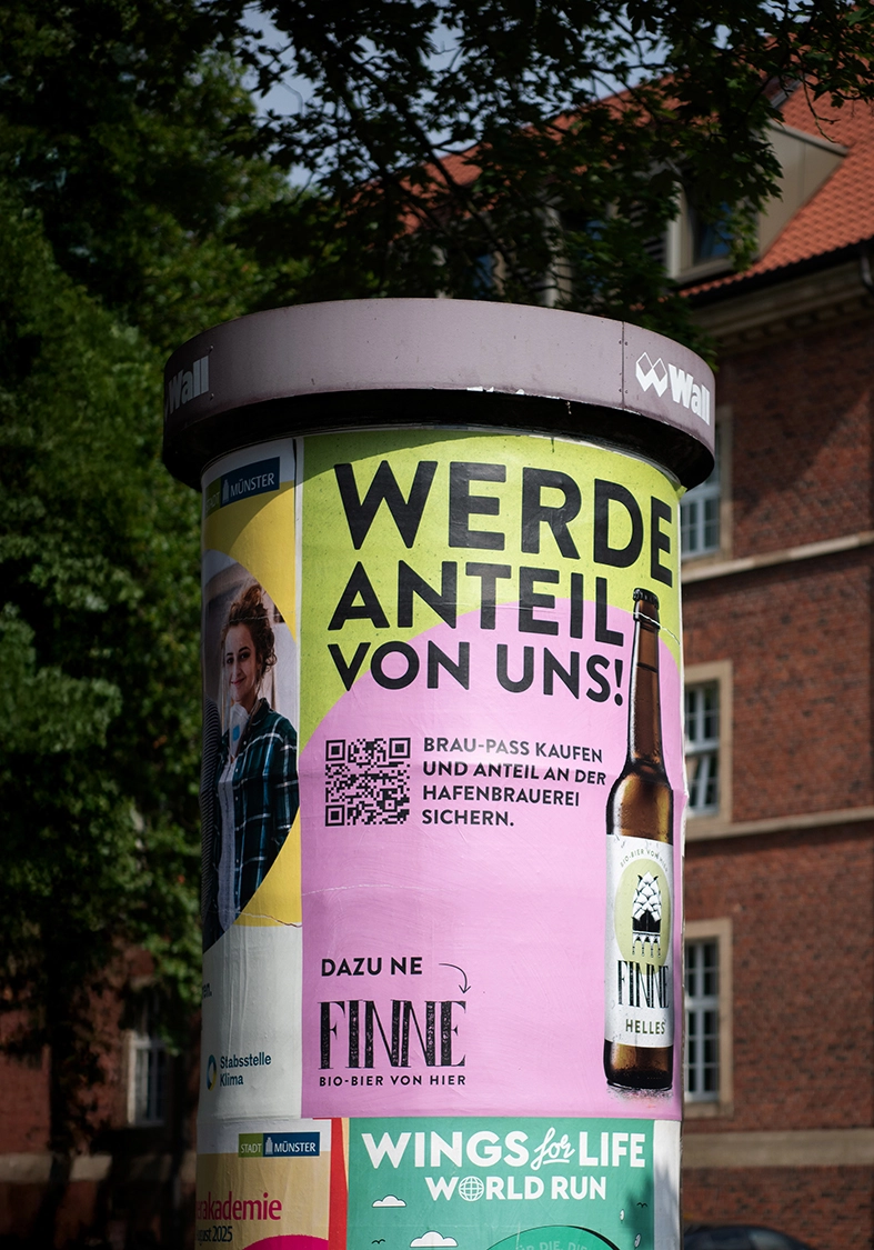 Eine mit bunten beklebte Litfasssäule im Sonnenschein. Auf der Litfasssäule ist ein Plakat der Brauerei Finne aus Münster mit einer Bierflasche zu sehen.
