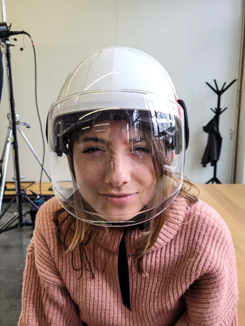 Eine weibliche Person in einem rosa Rollkragenpullover schaut lächelnd in die Kamera. Die Frau trägt einen weißen Schutzhelm mit einem transparenten Visier.