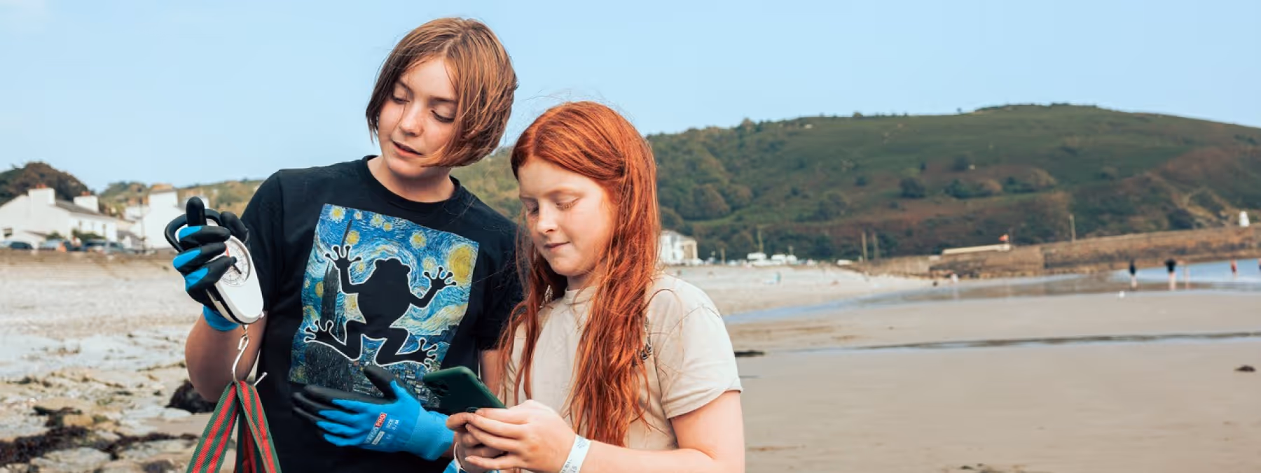 Two people on a beach - one weighing a bag of rubbish and the other recording the data on a phone and a big blue bag